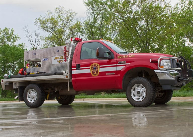 A red fire department brush truck with emergency lights is parked on wet pavement, surrounded by trees. The truck has equipment in the back and “Abcol VFD” is visible on the side.