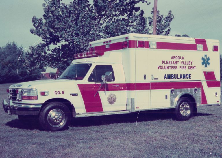 A red and white ambulance marked “Arcola Pleasant-Valley Volunteer Fire Dept.” is parked on grass near trees under a clear sky.