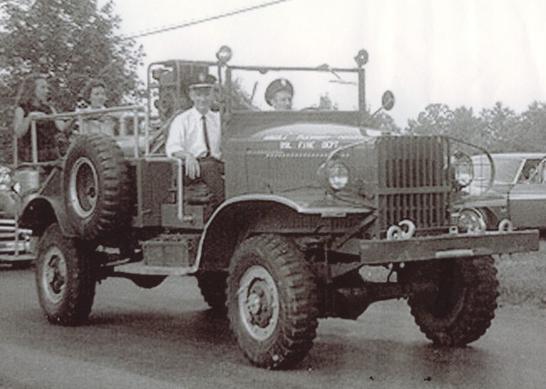 A vintage fire truck with several people riding on it is driving down a road. Trees and parked cars are visible in the background. The image appears to be from an earlier era, possibly mid-20th century.