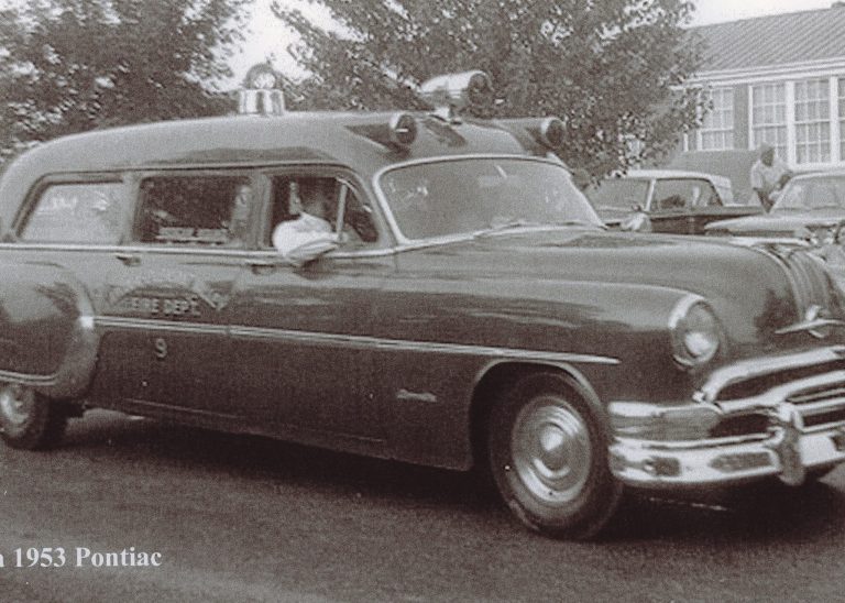 A vintage black-and-white photo shows a 1953 Pontiac ambulance labeled “FIRE DEPT” driving on a street, with a driver inside and classic cars and a building in the background.