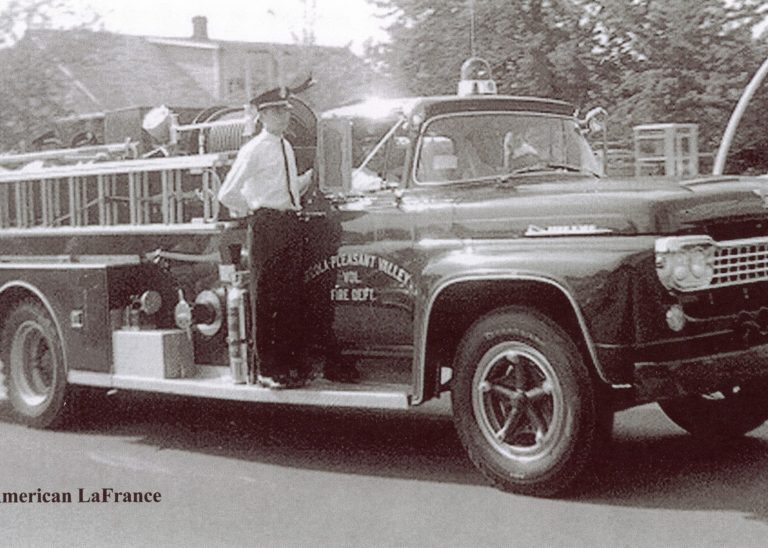 A vintage 1958 Ford American LaFrance fire truck drives down a street with a uniformed firefighter standing on the side. Trees and a building are visible in the background.