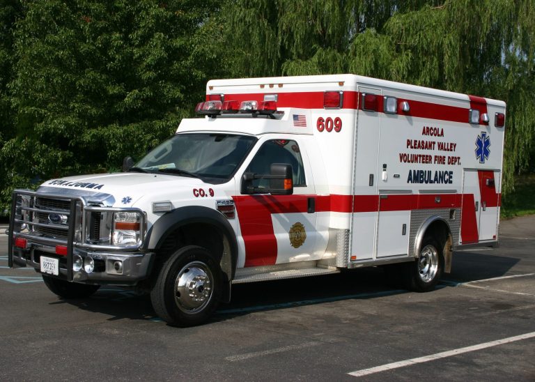 A white and red ambulance labeled “Ambulance” and “Arcola Pleasant Valley Volunteer Fire Dept.” is parked in a parking lot near green trees on a sunny day.