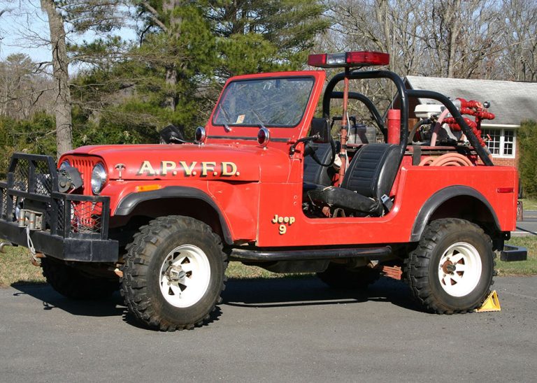 A vintage red Jeep with “A.P.V.F.D.” on the side, equipped with firefighting gear and hoses, parked on a paved surface with trees and a building in the background.