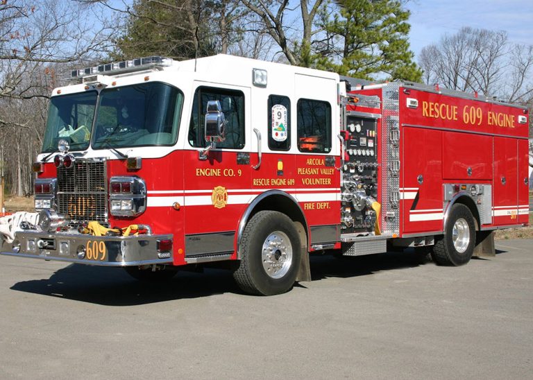 A large red fire engine labeled “Rescue 609 Engine” is parked outdoors on pavement, surrounded by trees and bushes. The vehicle displays emergency lights and reflective stripes, with various equipment compartments visible.