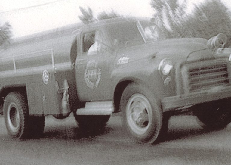 Black and white photo of a vintage fire truck in motion, with blurred background. The truck has a rounded front and emergency lights on top, and visible fire department logos on the door.