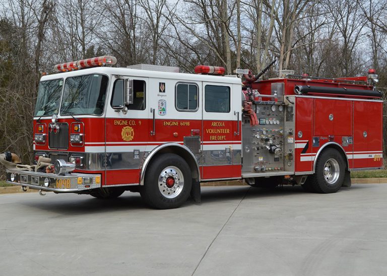 Red and white fire truck parked on a driveway with trees in the background. The truck belongs to a volunteer fire department and has hoses, dials, and emergency lights visible.