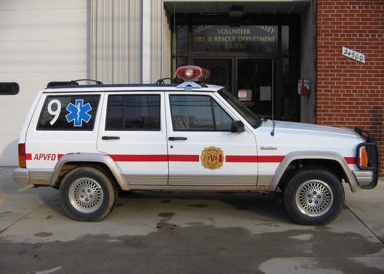 A white emergency vehicle with a red stripe, medical symbol, and fire department badge is parked outside a fire and rescue building.