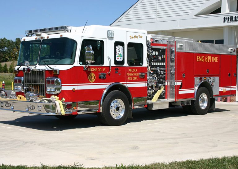 A bright red fire truck with white accents is parked outside a fire station on a sunny day. The truck has several hoses and equipment compartments visible on the side.