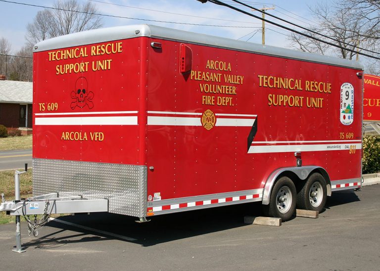 A bright red trailer labeled “Technical Rescue Support Unit” and “Arcola Pleasant Valley Volunteer Fire Dept.” is parked on pavement near a road, with trees and a brick building in the background.