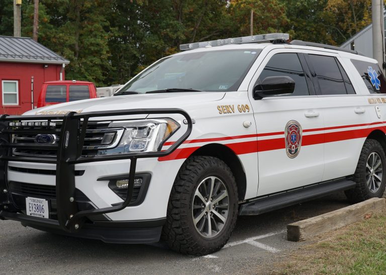 A white emergency response SUV with red stripes and a fire department emblem is parked next to a wooden barrier in a lot, with trees and a red building in the background.