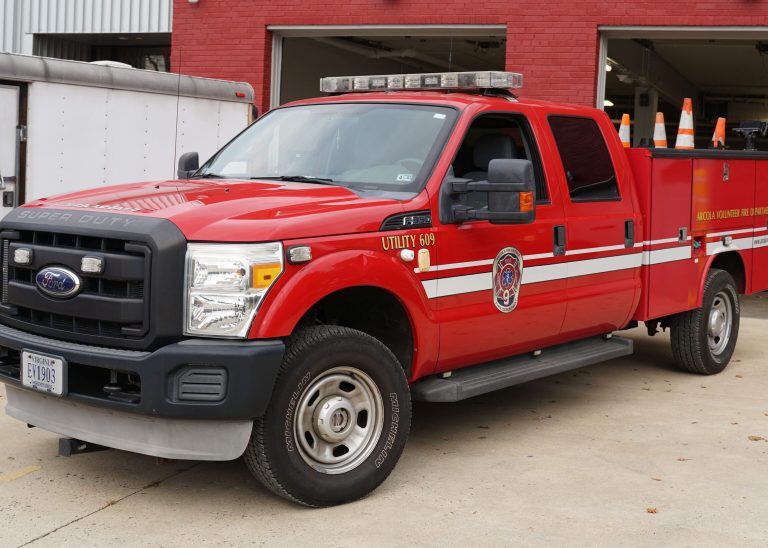 A red Ford Super Duty fire utility truck with emergency lights and orange traffic cones is parked outside a fire station with open garage doors.