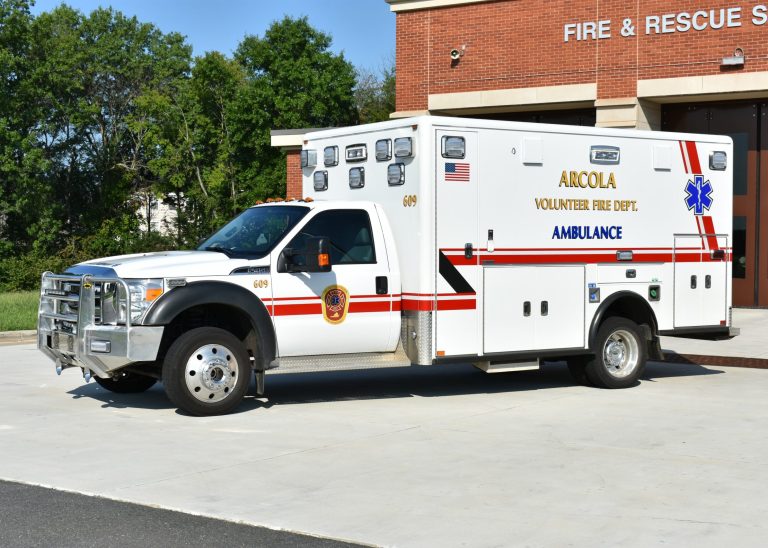 A white Arcola Volunteer Fire Dept. ambulance is parked outside a fire and rescue station on a sunny day.