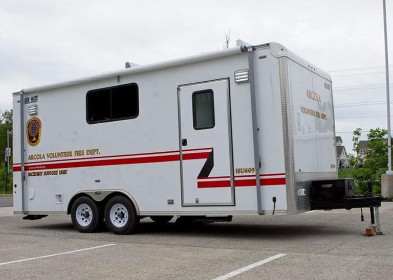 A white trailer labeled “ARCOLA VOLUNTEER FIRE DEPT. INCIDENT SUPPORT UNIT” is parked in a lot. It has emergency stripes, department logos, and two wheels on each side. Trees and pavement are visible in the background.