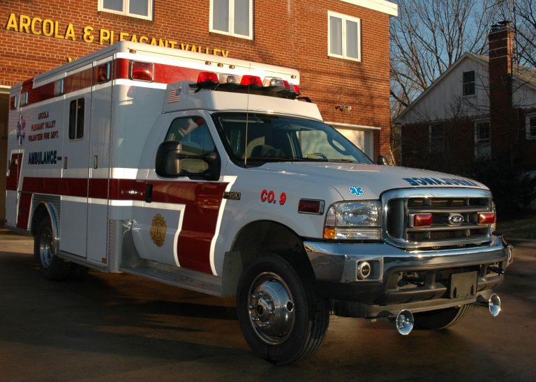 A white and red ambulance is parked outside a brick fire station, with the sun casting shadows. The vehicle has emergency lights on top and medical symbols on the side.