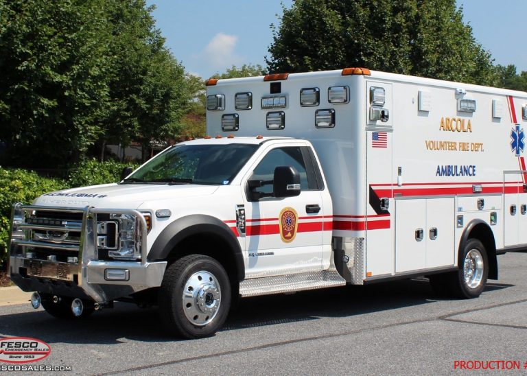 White ambulance truck with red stripes, labeled “ANOCLA VOLUNTEER FIRE DEPT.” parked outside on pavement, trees in the background, and emergency lights on top.
