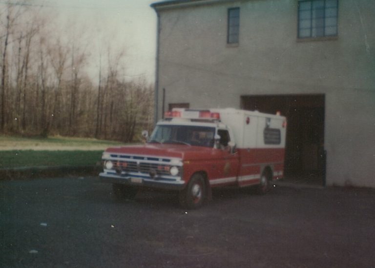 A vintage red and white ambulance is parked outside a beige building, with trees and grass visible in the background.