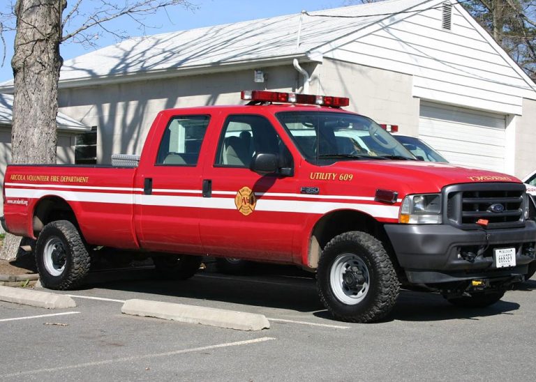 A red volunteer fire department utility truck with emergency lights is parked in a lot in front of a white building.